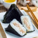 low angle shot of onigiri japanese rice triangle. One triangle is whole wrapped in seaweed. The other is cut in half so that you can see the inside. They are on a white plate on a bamboo mat.