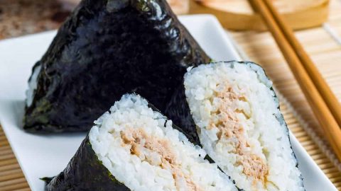 low angle shot of onigiri japanese rice triangle. One triangle is whole wrapped in seaweed. The other is cut in half so that you can see the inside. They are on a white plate on a bamboo mat.