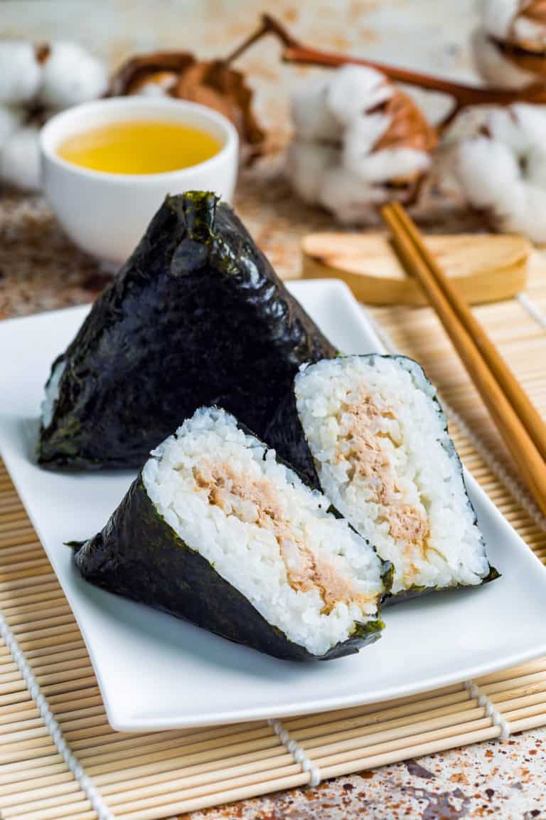 low angle shot of onigiri japanese rice triangle. One triangle is whole wrapped in seaweed. The other is cut in half so that you can see the inside. They are on a white plate on a bamboo mat.