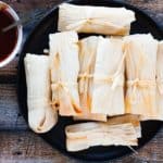 homemade tamales piled on a black plate with easy red chile sauce on the side