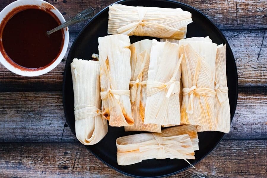 homemade tamales piled on a black plate with easy red chile sauce on the side