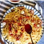 Top-down view of a plate of Burmese garlic noodles garnished with green onions and served with chopsticks. A hand holds a spoon with dark sauce above the garlic noodles, ready to drizzle.