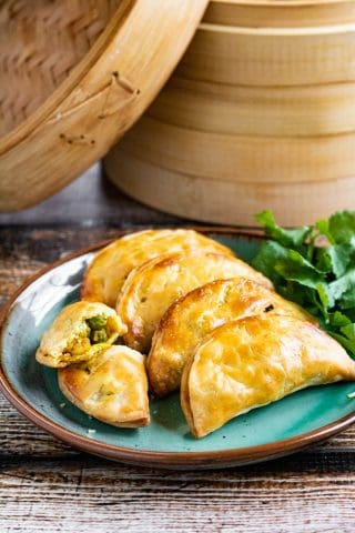 A plate with five golden-brown Thai curry puffs, one with a visible vegetable filling, sits next to fresh cilantro, with bamboo steamers in the background.