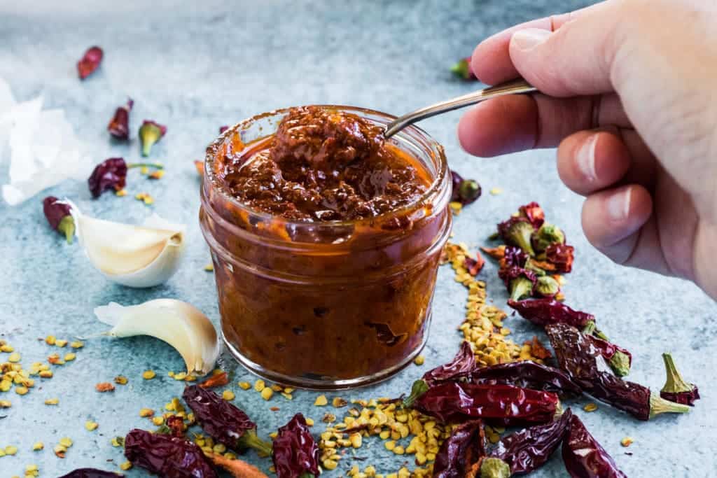 A hand is dipping a spoon into a jar of harissa sauce.