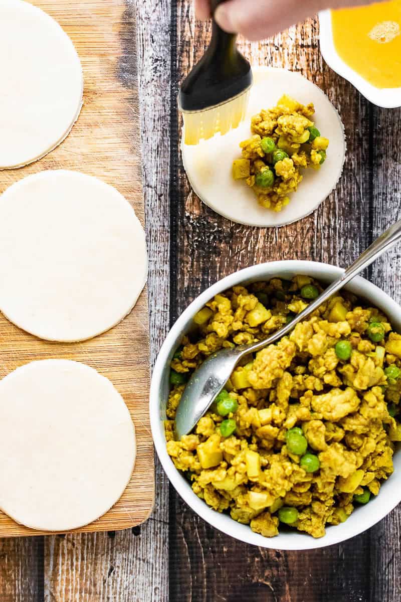 Round dough sheets on a wooden board and a bowl of curried filling with peas hint at the making of Thai curry puffs; a hand brushes liquid on a filled dough circle, preparing it to be folded and sealed.