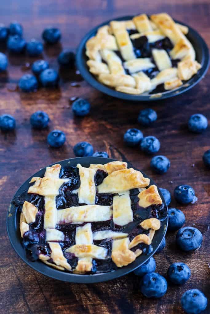 Two mini blueberry pies baked and ready to eat, with fresh blueberries scattered around them