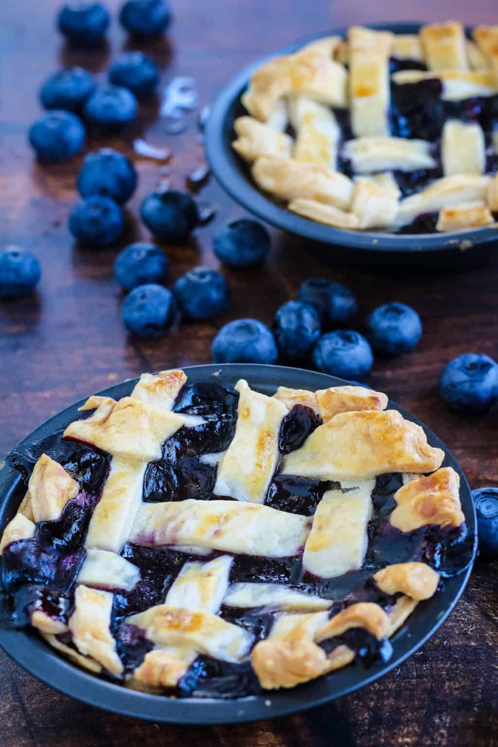 Mini blueberry pies shot from a low angle with fresh blueberries scattered around on a wood table.