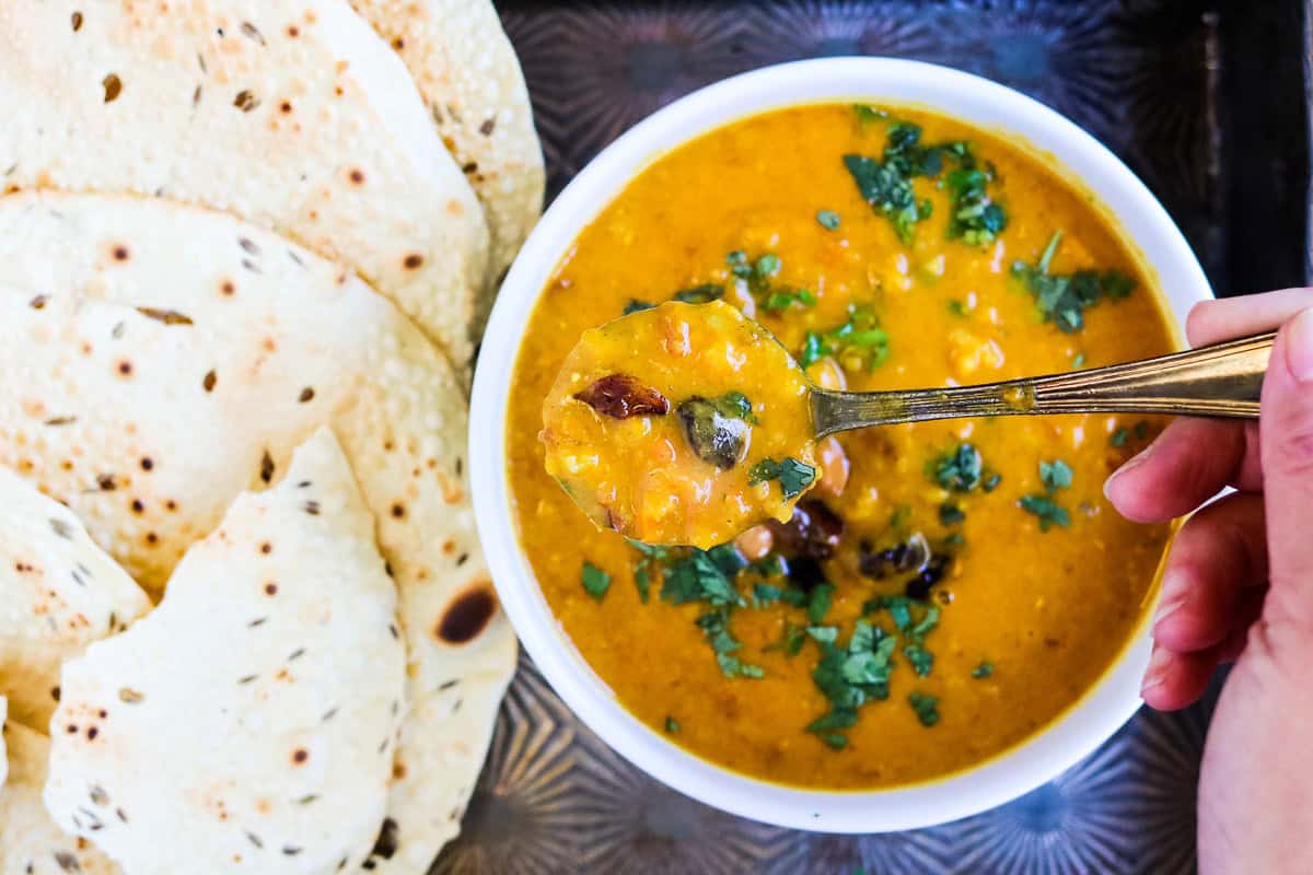 A hand holds a spoonful of yellow lentil mulligatawny soup with herbs above a bowl, next to pieces of flatbread on a tray.