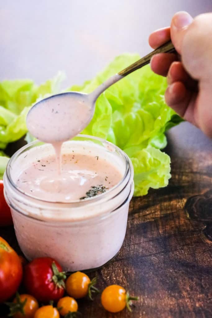 A spoonful of creamy sriracha dressing being lifted out of a jar with lettuce and tomatoes on the side