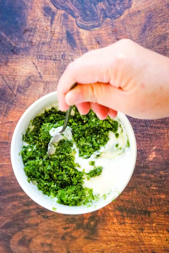 minced cilantro, ginger, chilies, and garlic being stirred into yogurt in a bowl.
