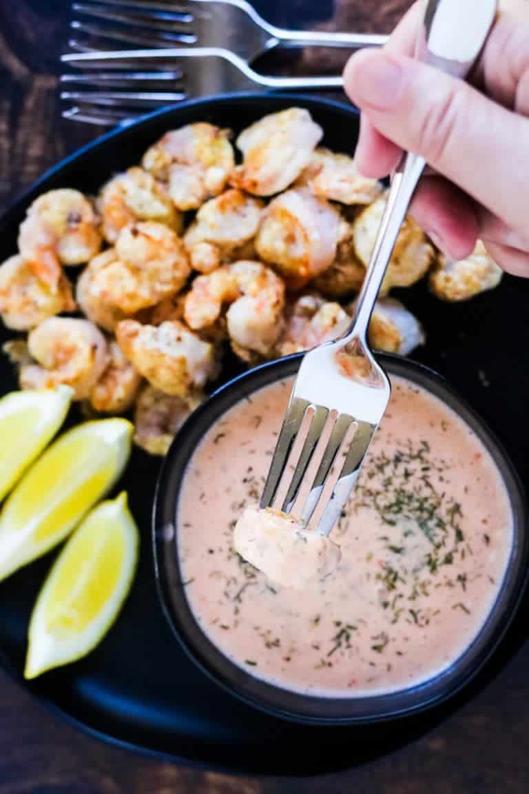 overhead shot of air fryer fried bang bang shrimp with sauce and lemon wedges. There is a hand holding a fork with one shrimp on it that has been dipped in the sauce.