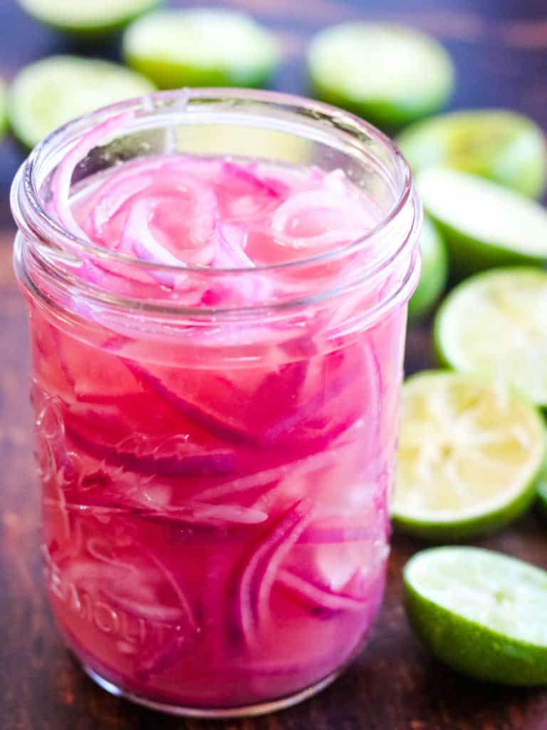low angle shot of a mason jar filled with pickled red onions. There are lots of halved limes in the background