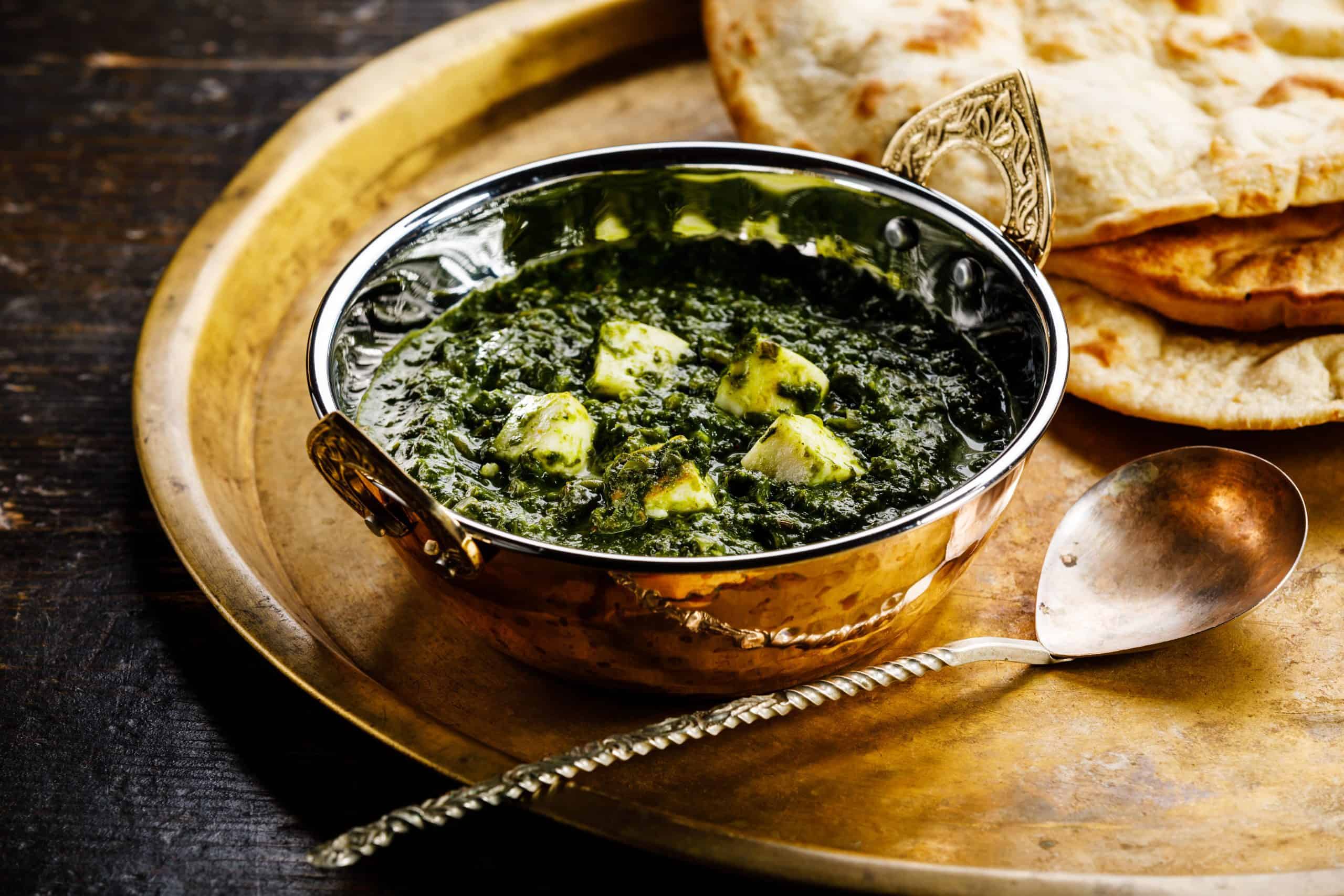palak paneer in a metal bowl on a metal tray with a spoon and naan bread, shot from a low angle