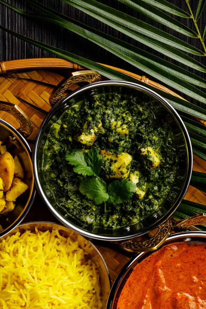 overhead shot of Indian cheese and spinach curry in a metal bowl. On the side are a bowl of yellow rice, a bowl of masala curry, and a palm frond