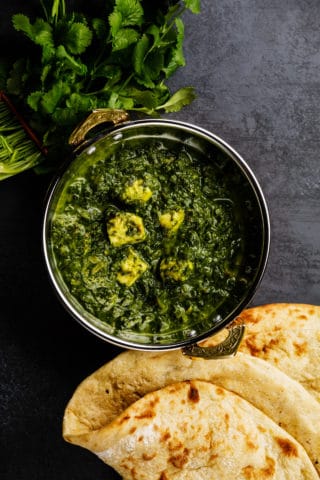 Overhead shot of palak paneer in a metal bowl on a gray stone counter. Next to the bowl of paneer, there is a bunch of cilantro and two pieces of naan bread