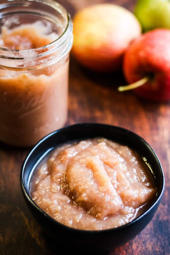 low-angle vertical shot of homemade instant pot applesauce in a black bowl with a jar of applesauce and some apples in the background
