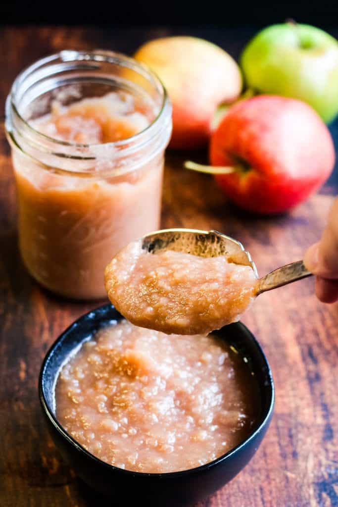 low-angle vertical shot of homemade instant pot applesauce in a black bowl with a spoonful being lifted a jar of applesauce and some apples in the background