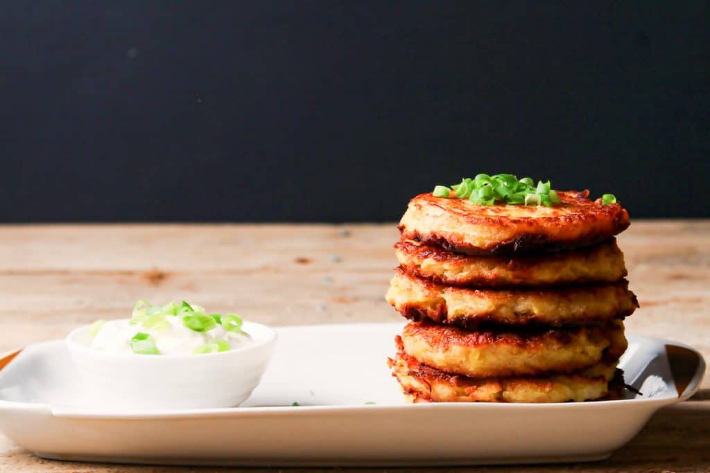 Low angle photo of a stack of 5 potato latkes on a white rectangular plate. There is a bowl of sour cream on the plate and both the sour cream and potato latkes are garnished with sliced green onions.