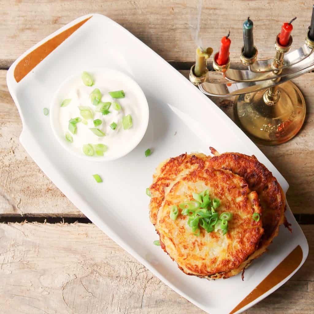 Overhead photo of a stack of 5 potato latkes on a white rectangular plate. There is a bowl of sour cream on the plate and both the sour cream and potato latkes are garnished with sliced green onions. There is a menorah next to teh plate with blue and red candles in it