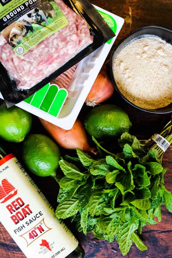 Ingredients for thai larb including ground meat, roasted rice powder, limes, shallots, fish sauce, and fresh mint