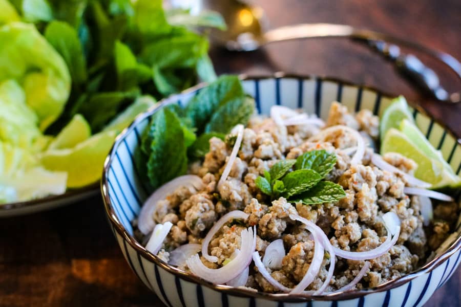 Landscape shot of thai larb in a blue and white serving bowl with fresh mint, lime wedges, sliced shallots, and lettuce leaves