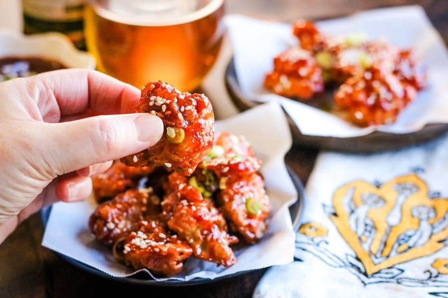 Korean fried chicken on a plate with a beer and someone holding a piece of the chicken in their fingers