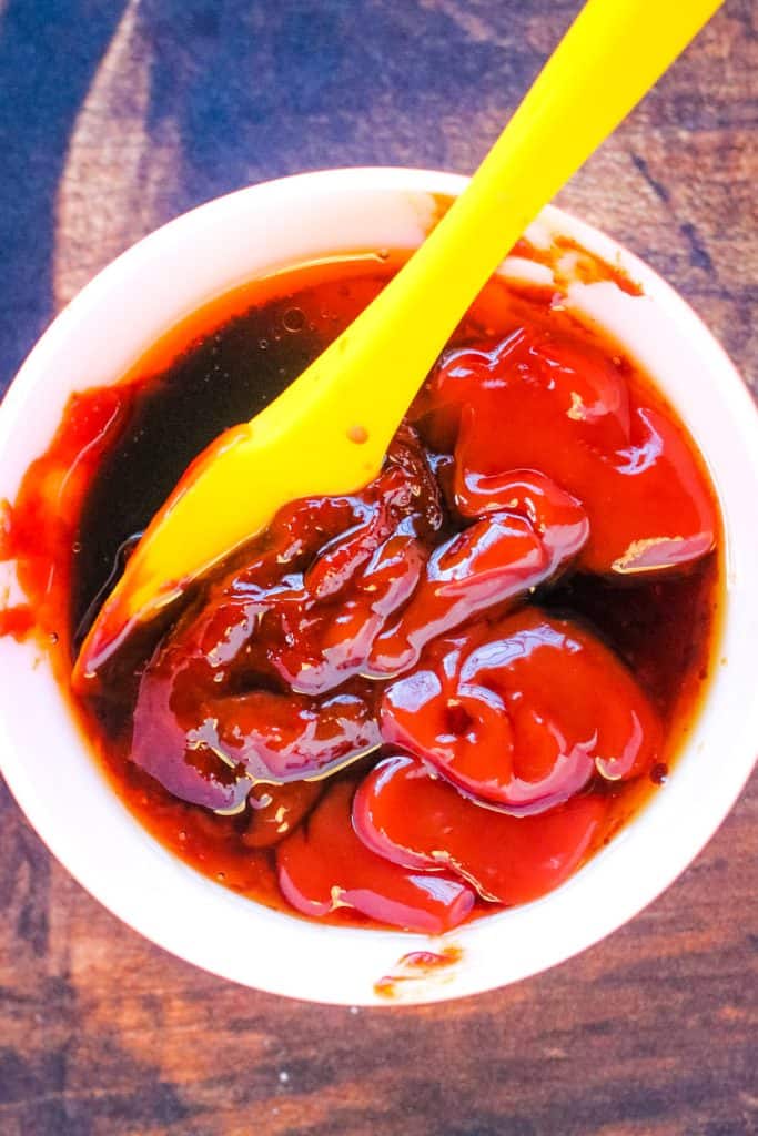 A white cup brimming with dark red sauce, perhaps for pairing with air fryer Korean fried chicken, features a yellow plastic spoon resting atop its wooden surface.