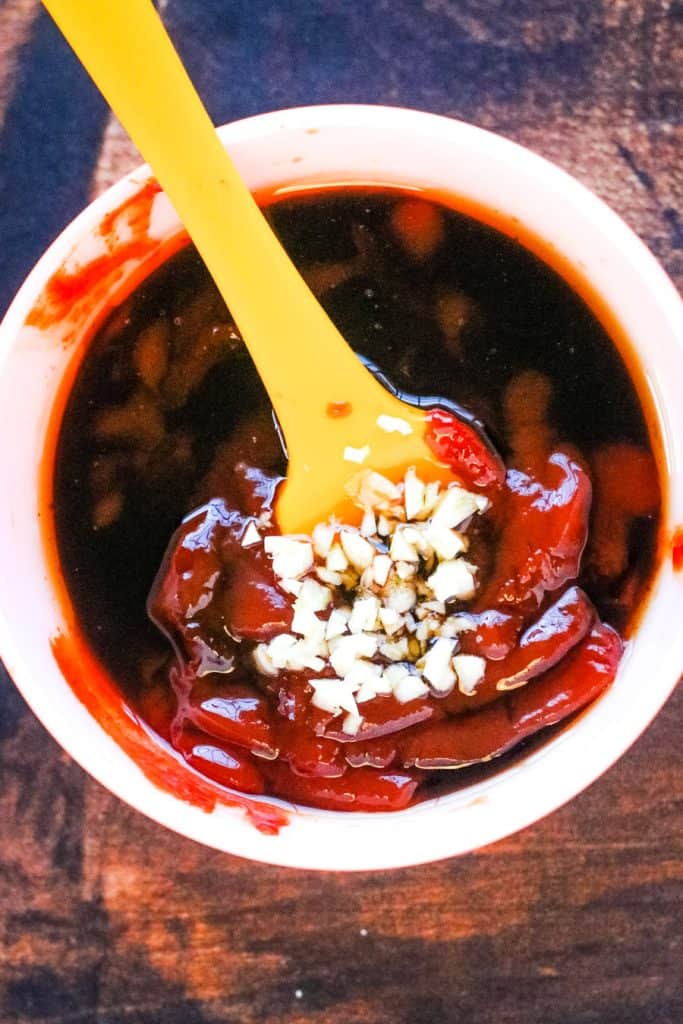 A container filled with a dark sauce, topped with chopped white garlic and a swirl of red sauce, reminiscent of the flavors one might find in air fryer Korean fried chicken, with a yellow spoon resting inside.