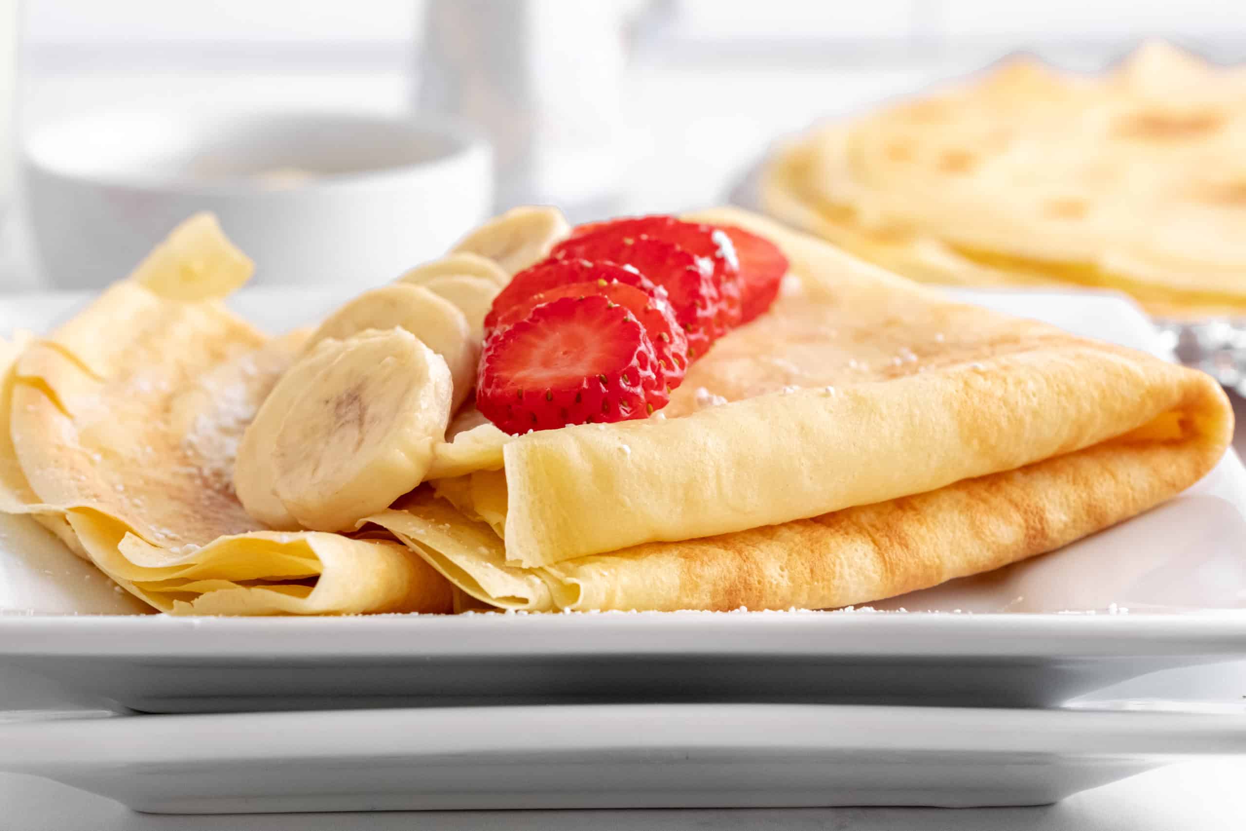 Low angle shot of a crepe folded up on a white plate with sliced banana and strawberry on top.