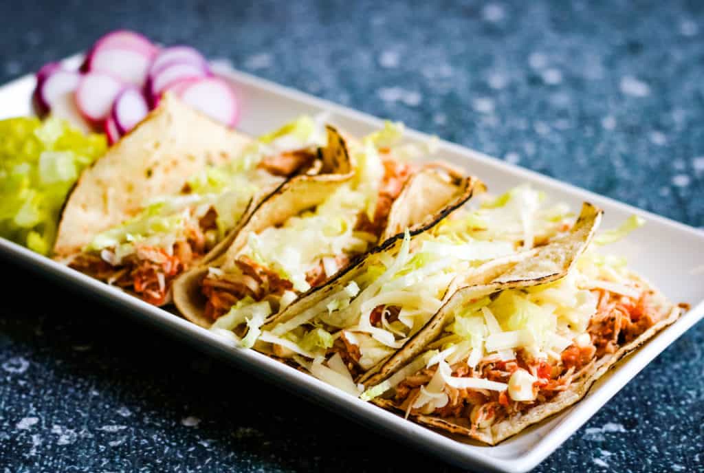 Low angle shot of 4 shredded chicken tacos in soft corn tortillas on a rectangular white plate. There is a pile of shredded lettuce and some sliced radishes on the plate.
