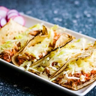 Low angle shot of 4 shredded chicken tacos in soft corn tortillas on a rectangular white plate. There is a pile of shredded lettuce and some sliced radishes on the plate.