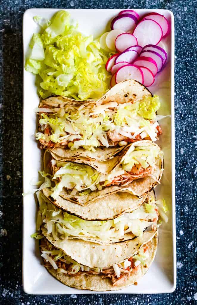 overhead shot of 4 shredded chicken tacos with shredded cheese, lettuce, and sliced radishes on a rectangular white plate.