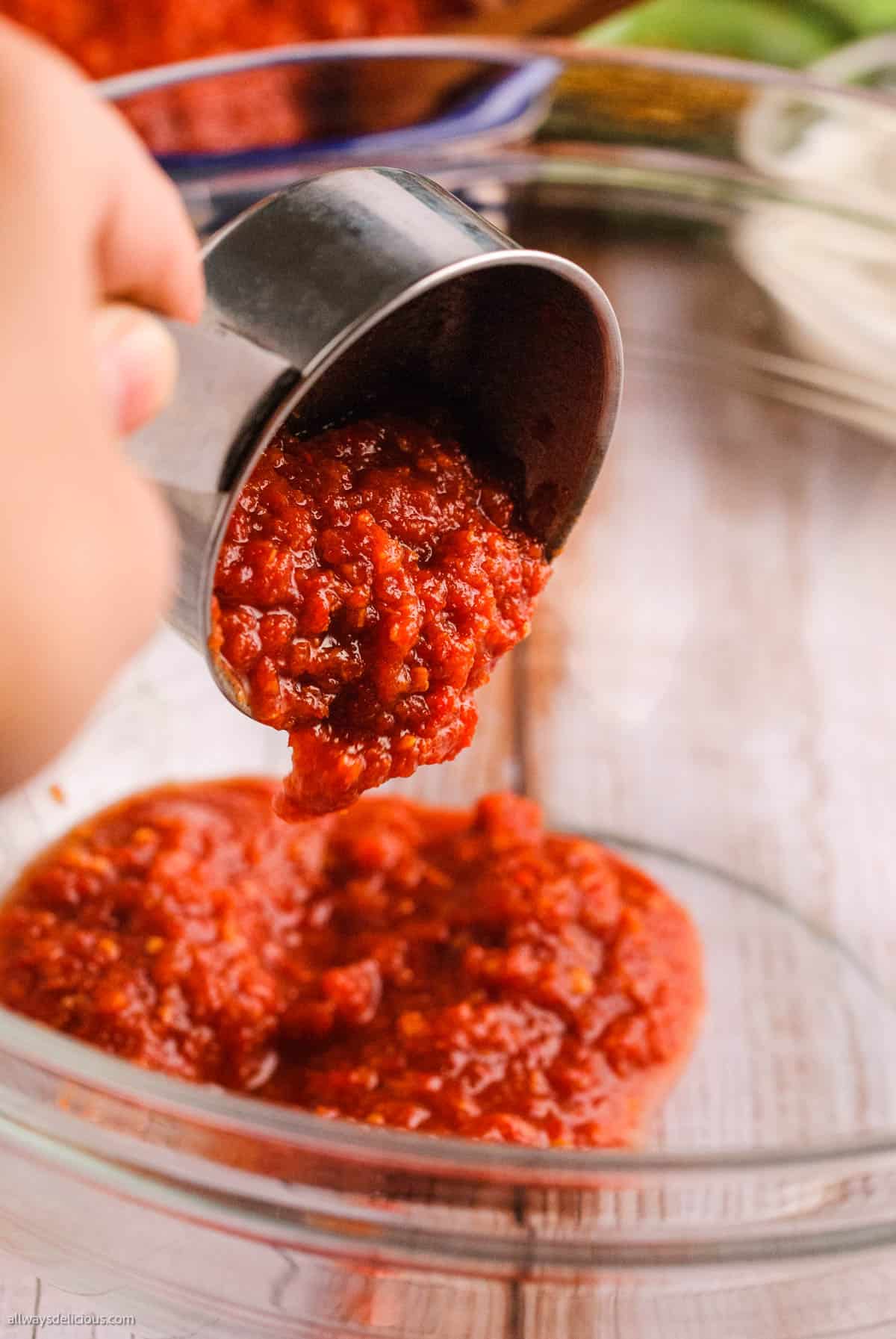 low angle shot of a hand pouring harissa paste from a measuring cup into a bowl