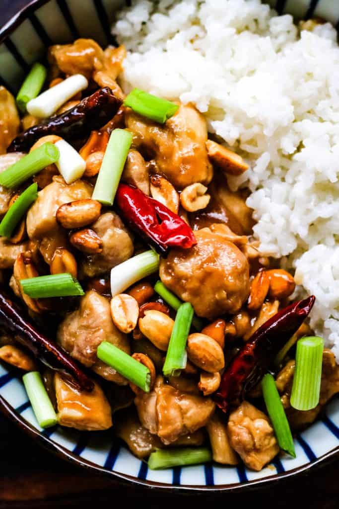 Overhead photo of kung pao chicken in a blue and white striped bowl. The chicken is cooked with red chiles, scallions, and peanuts and served alongside white rice.
