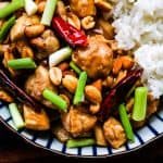 kung pao chicken in a blue and white striped bowl. There are chunks of chicken stir fried with chilies, peanuts, scallions and a savory sauce. There is cooked white rice on the side.