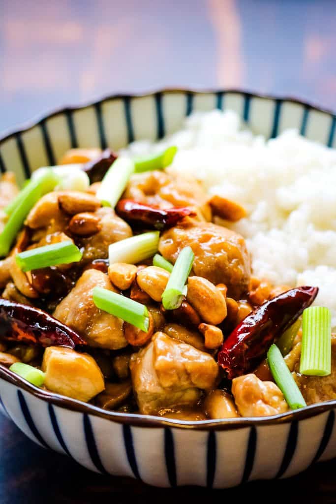 vertical photo of kung pao chicken in a blue and white striped bowl. The chicken is cooked with red chiles, scallions, and peanuts and served alongside white rice.