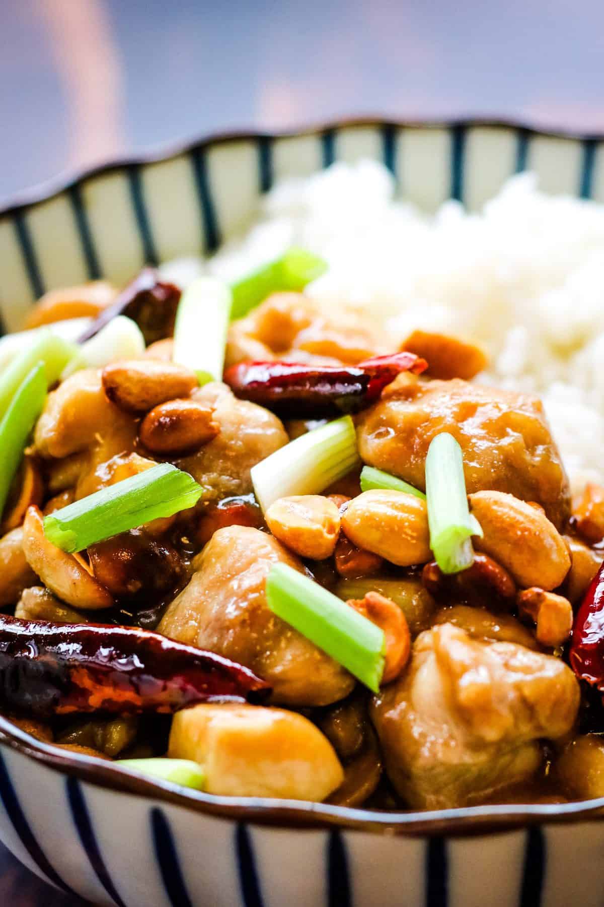 vertical photo of kung pao chicken in a blue and white striped bowl. The chicken is cooked with red chiles, scallions, and peanuts and served alongside white rice.