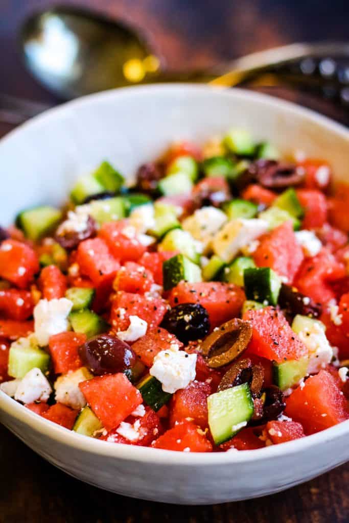 watermelon salad with cucumbers, feta cheese, and olives in a white bowl, shot from a low angle with silver salad servers in the background.