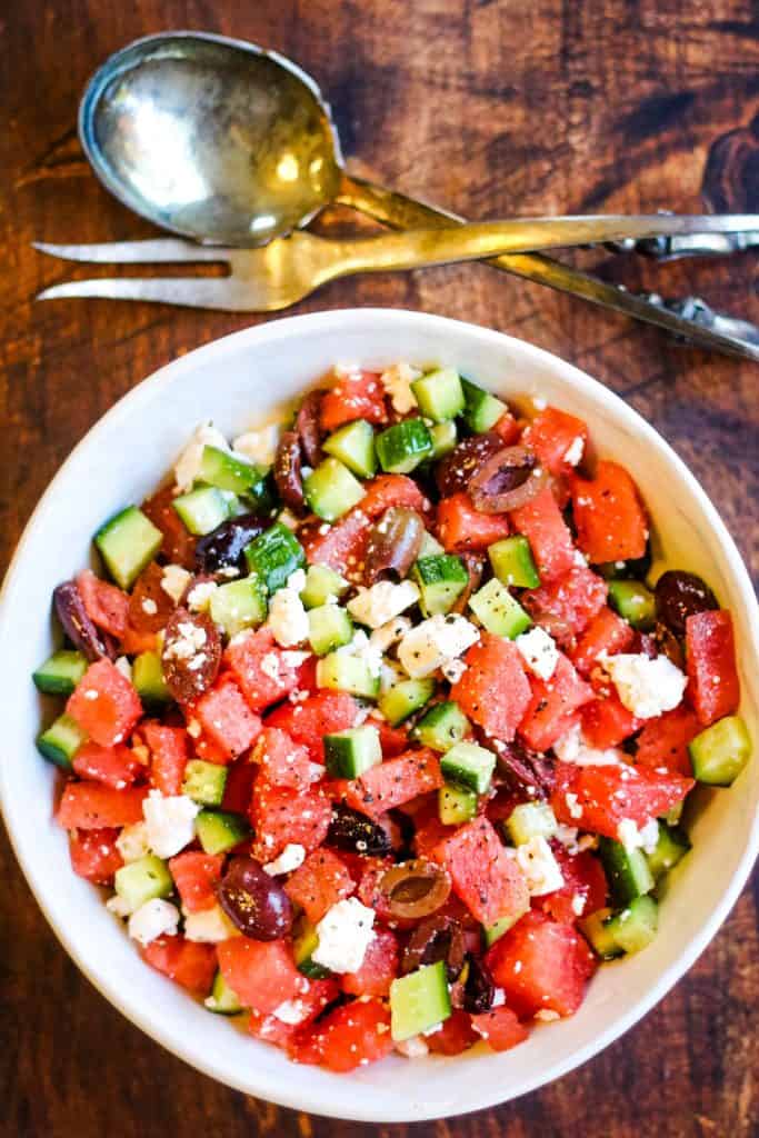 overhead shot of watermelon salad with cucumbers, feta cheese, and olives in a white bowl with silver salad servers on the table above the bowl.