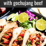 Pinterest pin for korean tacos with gochujang beef. The image is a low angle photo of corn tortillas filled with korean beef. There are lime halves and shredded purple cabbage on the side.