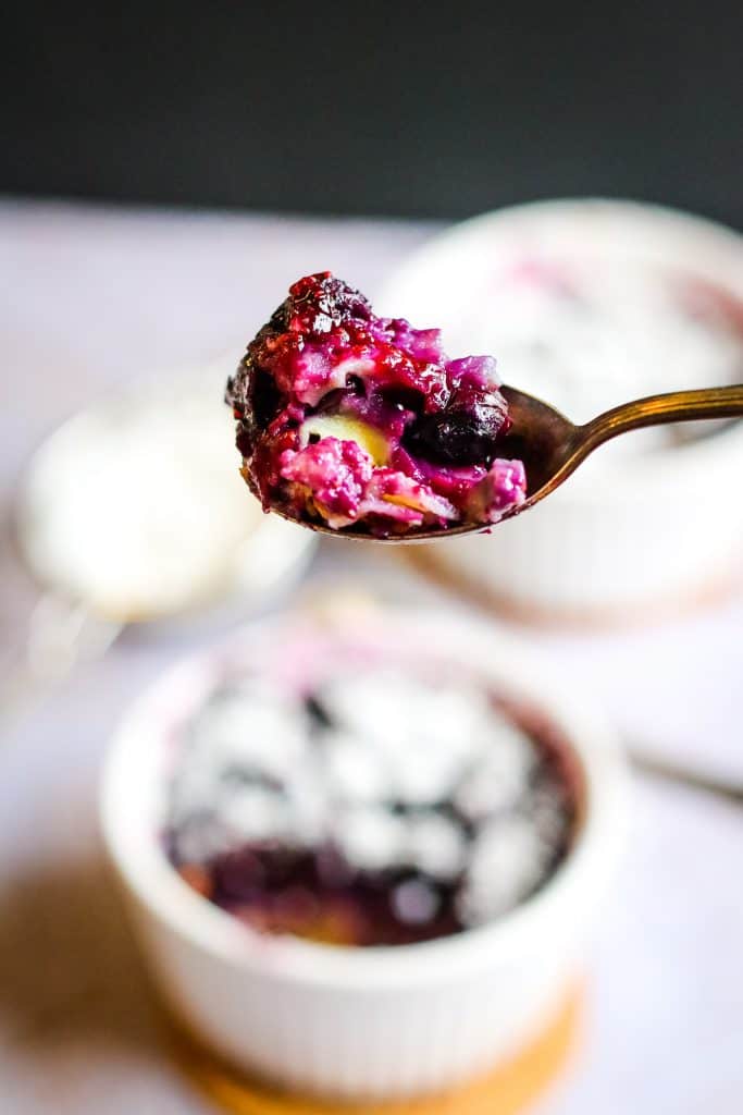 A low angle shot of blueberry clafoutis on a spoon. In the background there is a blurred out ramekin of blueberry clafoutis topped with powdered sugar.