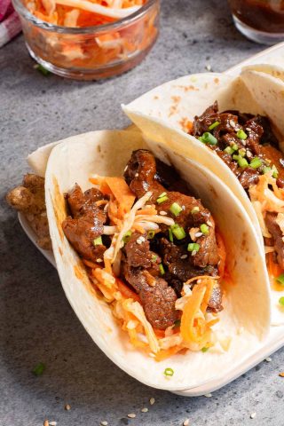 Close-up of two soft Korean taco shells filled with beef, shredded vegetables, and garnished with chopped chives, on a gray surface.