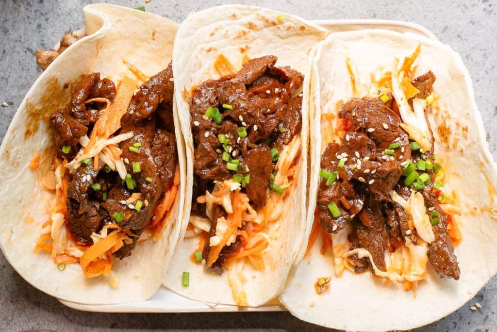 Three tortillas filled with seasoned beef, shredded vegetables, and topped with sesame seeds and chopped green onions create the perfect Korean taco experience, all elegantly arranged on a rectangular white plate.