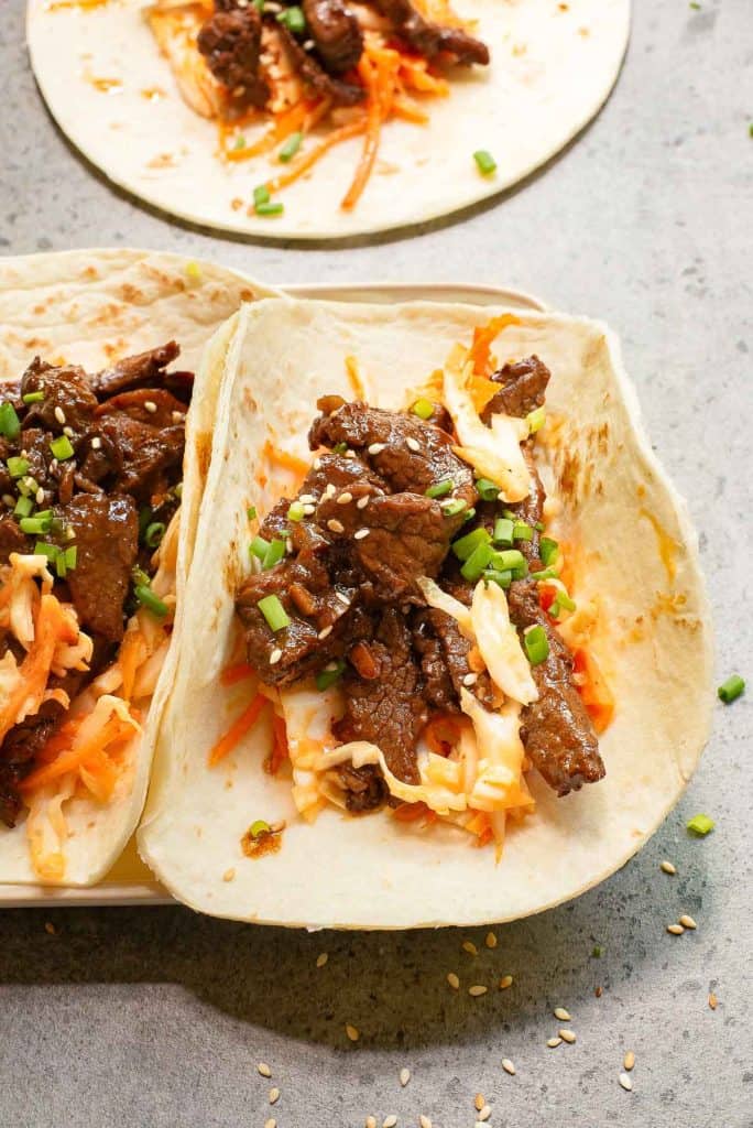 A Korean taco with seasoned beef, shredded vegetables, and green onions on a soft tortilla beckons your taste buds with its savory allure, while another taco tantalizingly peeks from the background.
