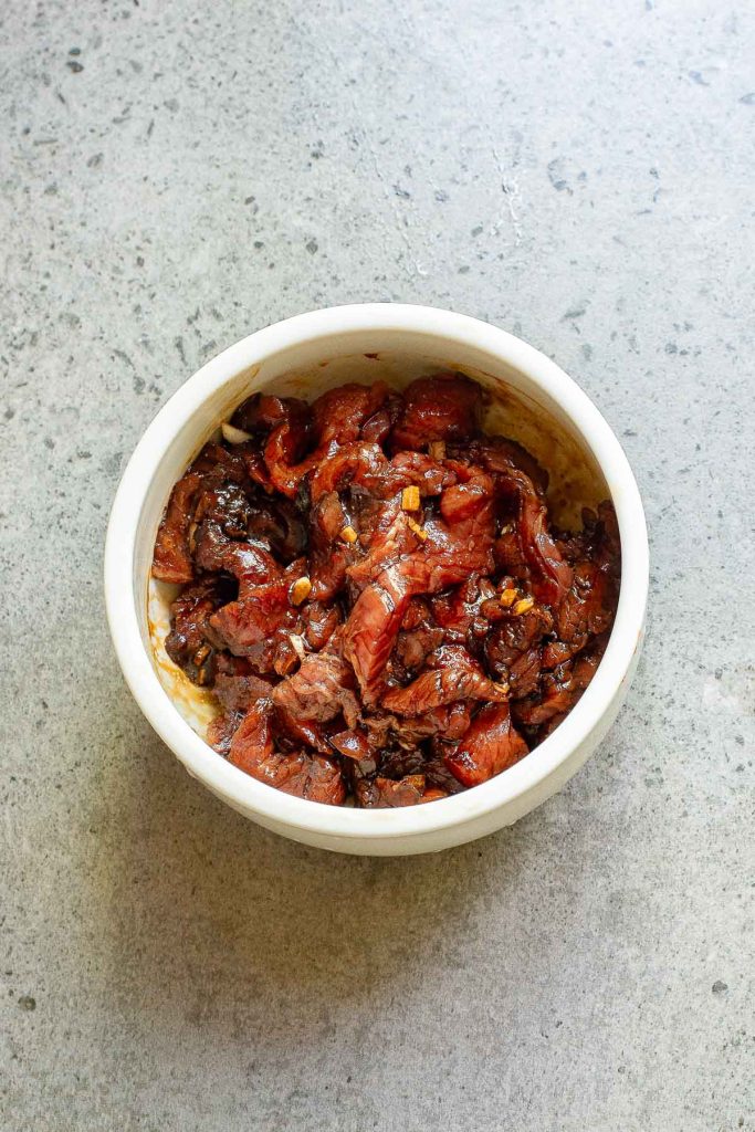 A white bowl filled with marinated sliced beef, perfect for Korean tacos, sits on a gray countertop.