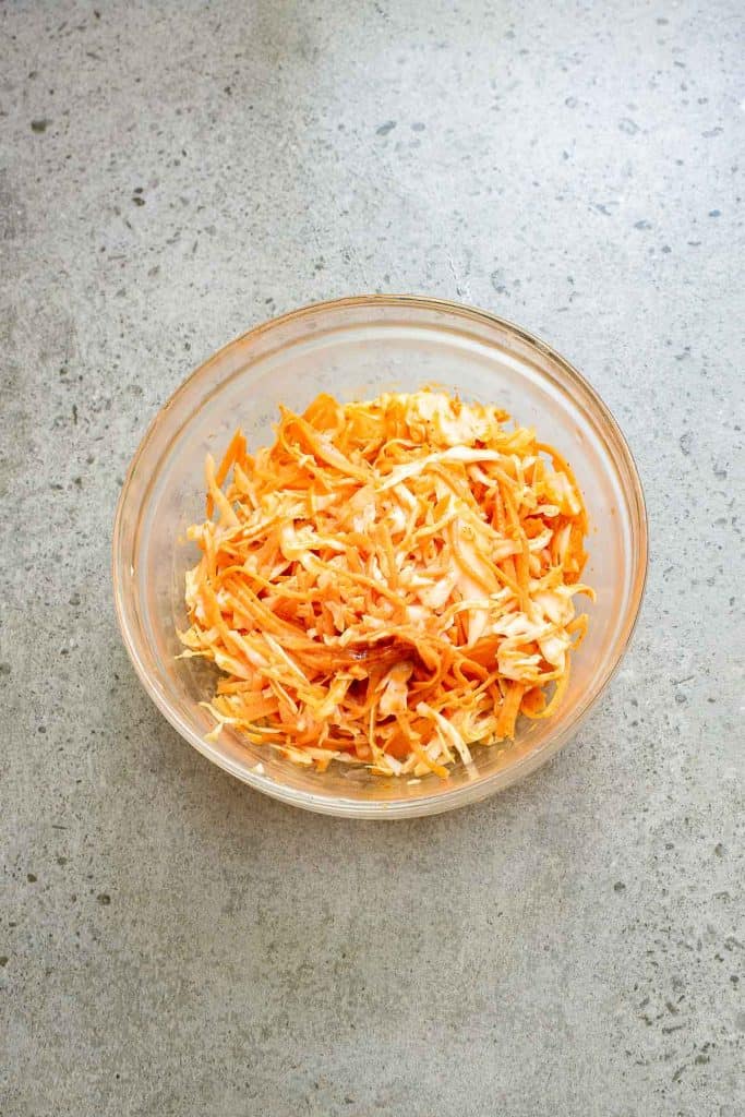 A clear bowl of coleslaw with shredded cabbage and carrots sits on a speckled gray countertop, perfectly complementing a feast of flavorful Korean tacos.