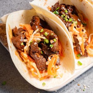 Two Korean tacos filled with marinated beef, shredded cabbage, and diced green onions, garnished with sesame seeds.