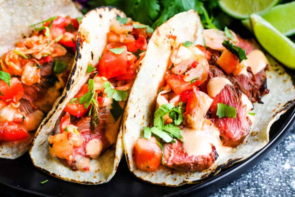Korean-style steak tacos on a black plate with tomatoes and limes.