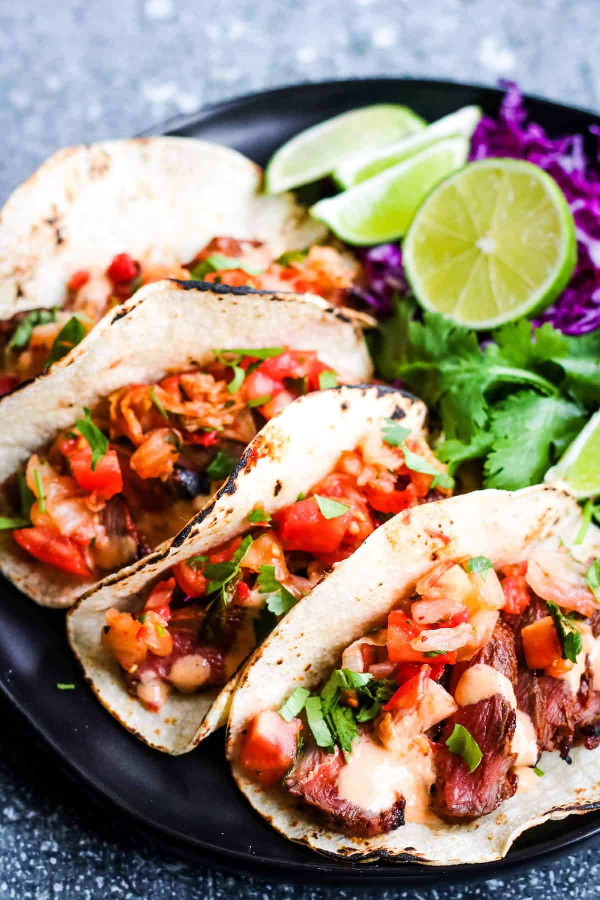 Vertical shot shot of 4 korean tacos: corn tortillas filled with sliced steak, kimchi and pico de gallo salsa, and chopped cilantro. In the background are lime wedges, sliced red cabbage, cilantro leaves, and corn tortillas.