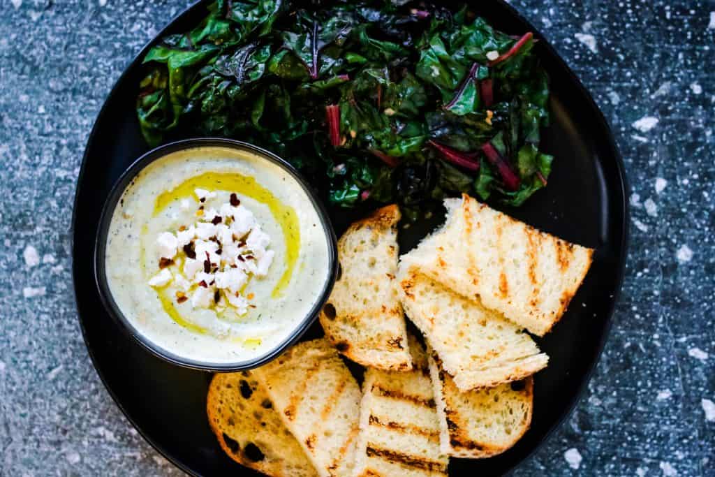 overhead shot of a black platter with a bowl of whipped feta spread, sauteed chard, and grilled sourdough bread
